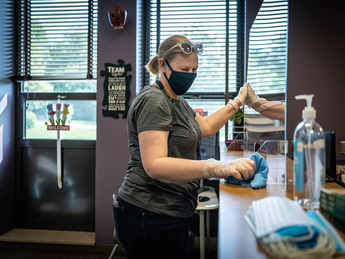 A worker in gloves and a mask wiping down a desk surface with disinfectant
