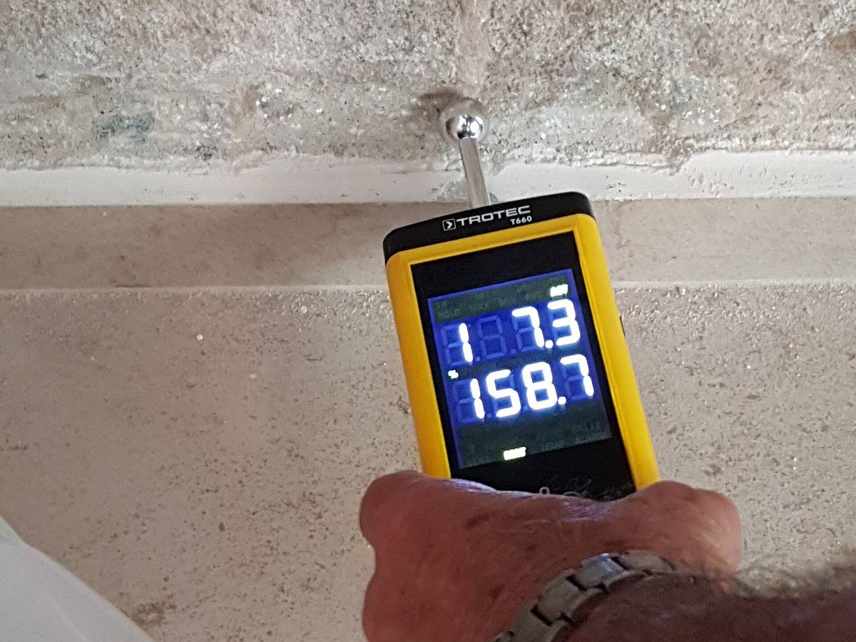 A hand holding a moisture meter against a wall, its display reading moisture hidden in the material