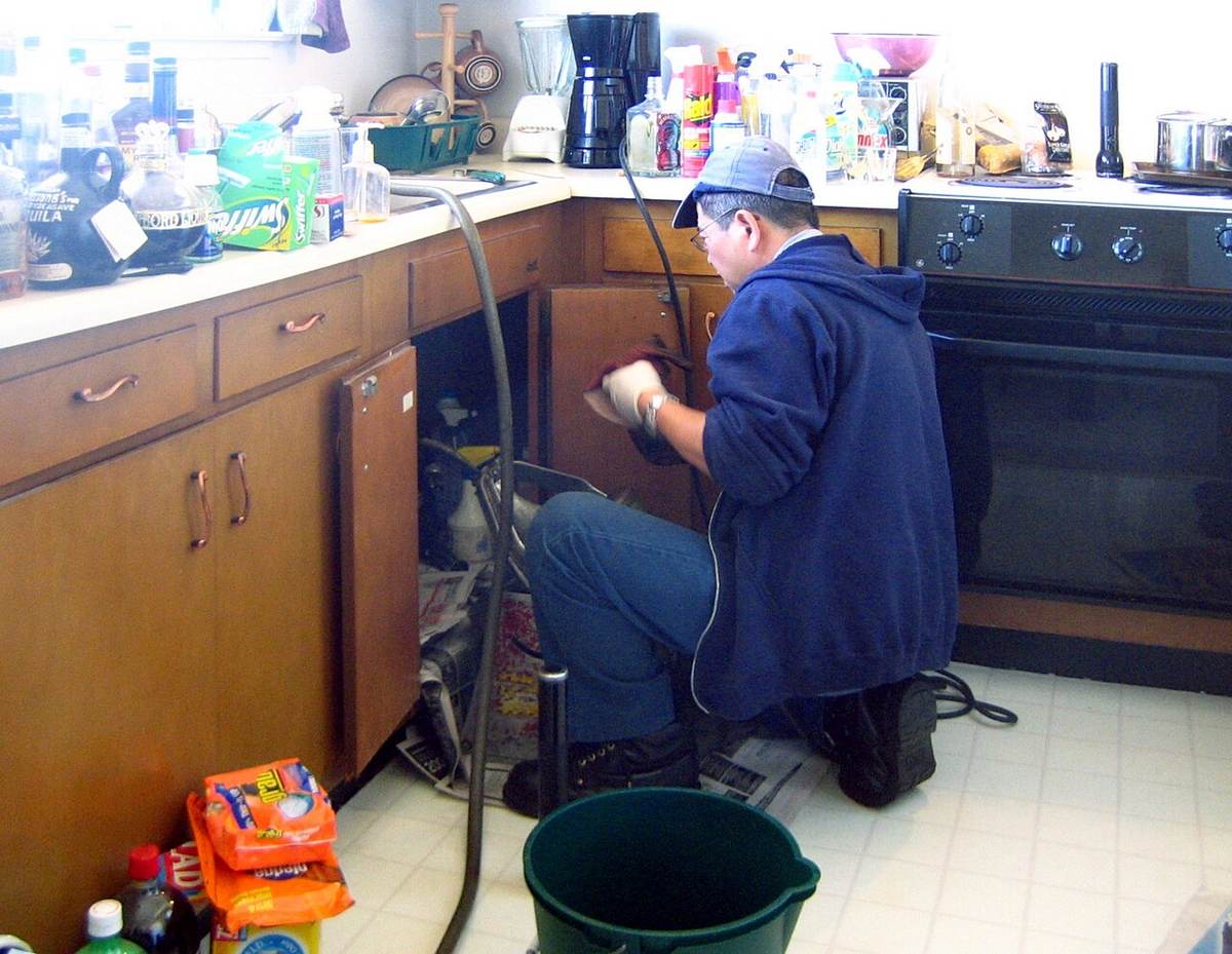 Plumber repairing under-sink plumbing in a Tulsa kitchen while a bucket catches water from a burst pipe