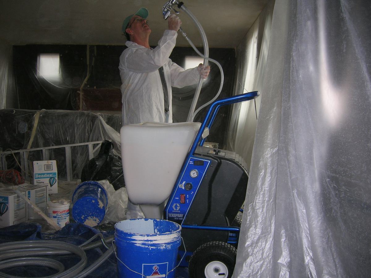 A worker spray painting a repaired ceiling, plastic sheeting protecting the room
