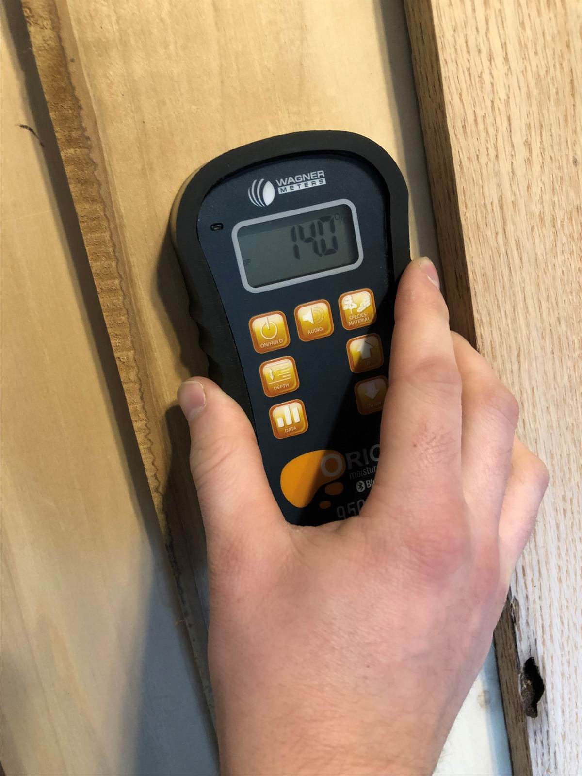 A hand holding a pin moisture meter against wood, showing a reading