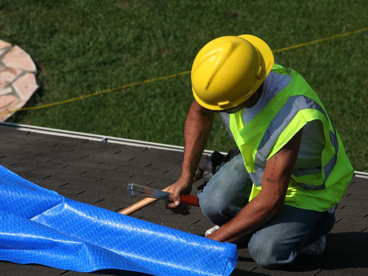 Worker nailing down a blue tarp over a damaged roof to stop rain getting in