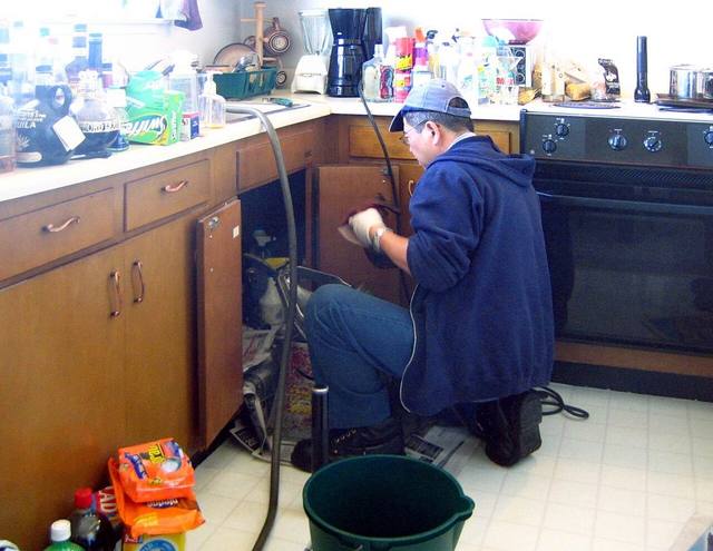 Plumber repairing under-sink plumbing in a Tulsa kitchen while a bucket catches water from a burst pipe