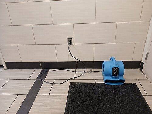 A blue air mover fan drying a tiled floor against a wall